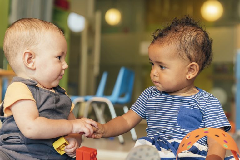 Two babies playing with colorful plastic toys on the floor.