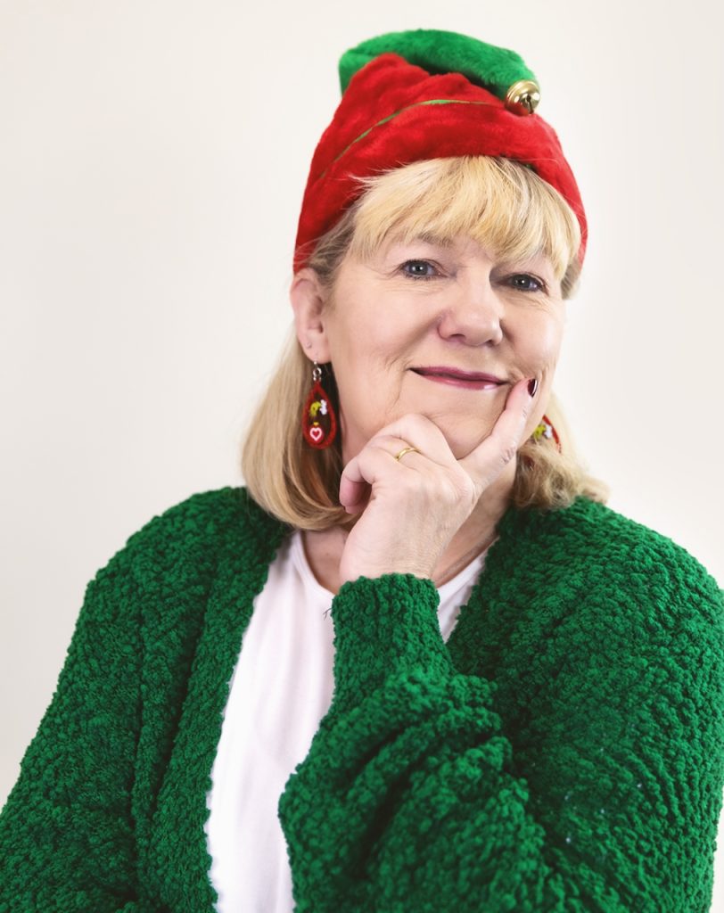Suzy wearing a white shirt and a green sweater, a red and green elf had with a jingle bell, and Grinch earrings. She's smiling and has her hand to her face with her index finger extended beside her smile.