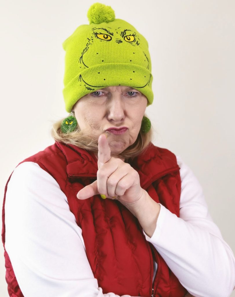 Suzy wearing a white shirt and red puffy vest, a green knit Grinch hat, Grinch-green nail polish and Grinch earrings. She's shaking her finger and scowling (in a fun way) at the camera.