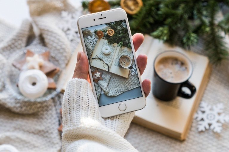 Cell phone held by a hand of person wearing soft cream-colored sweater. In the background is a mug of warm liquid, probably coffee. On the phone screen is a book, a pair of eyeglasses, a mug of foamy coffee, and greenery with dried fruit and stars made of something like clay.