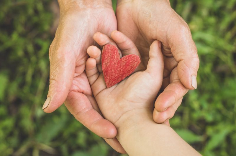An elderly person's upturned hands holding a child's hand that's holding a red heart.