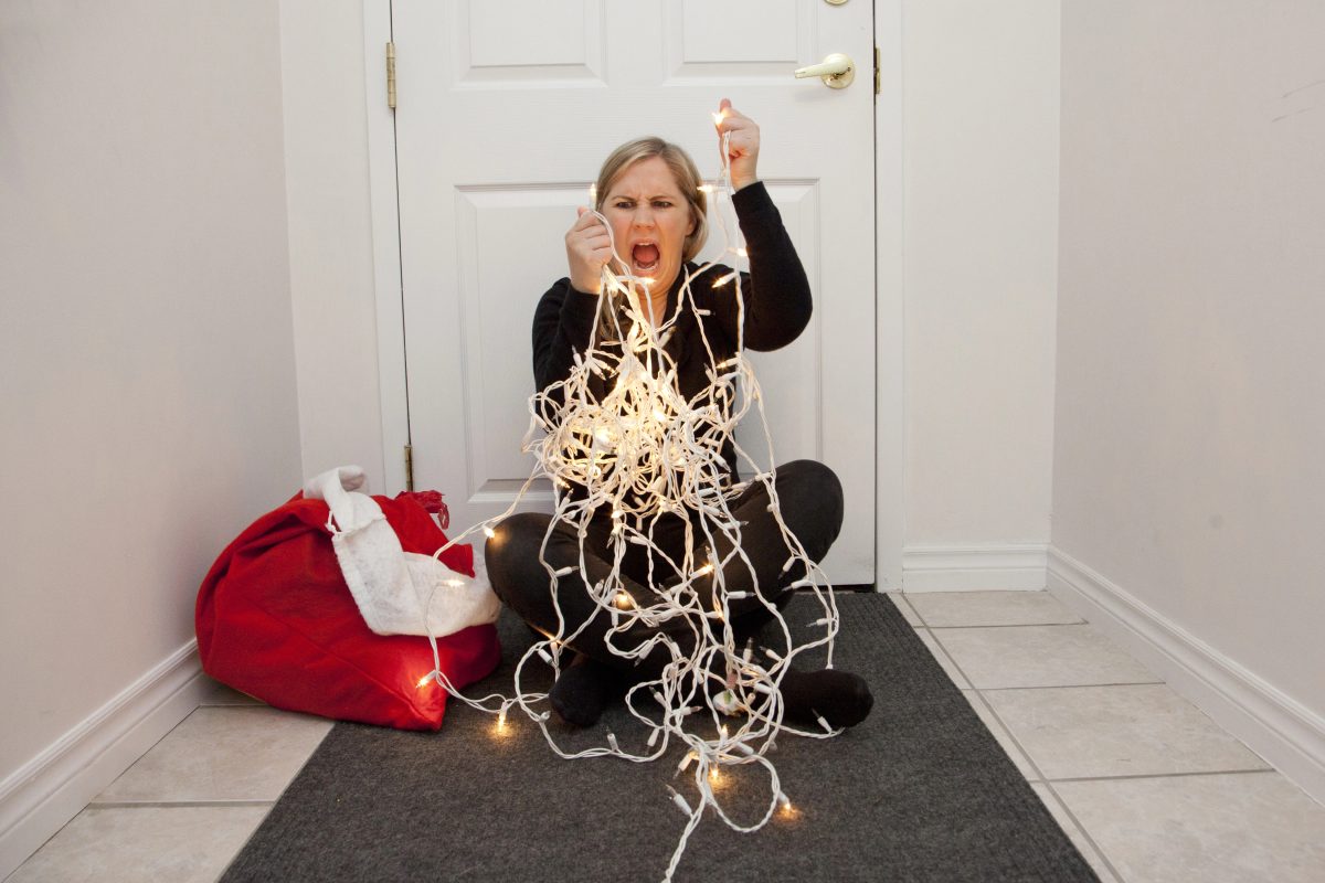 Frustrated woman sitting on floor with tangled mess of white Christmas lights. Red and white bag sits on the floor next to her.