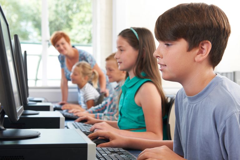Kids sitting at computers with woman looking down the row at them.