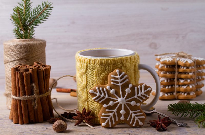 Warm scene with mug wrapped in yellow knitted cozy surrounded by cinnamon sticks and gingerbread cookies tied with natural twine. Nuts and spices are interspersed throughout the scene.