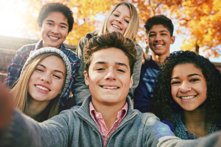 Multicultural group of five teenagers taking a selfie and smiling in front of an autumn-leaved tree.