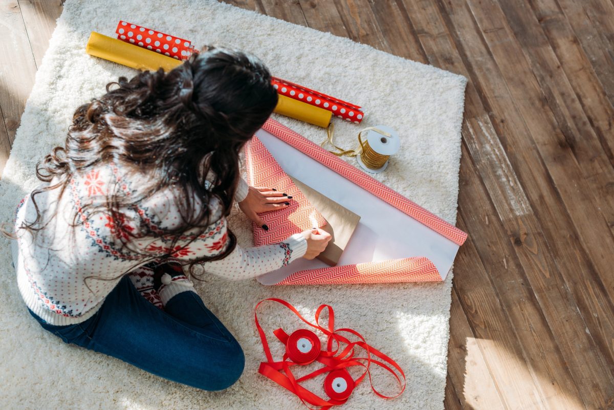 Overhead shot of a woman with long, dark hair wrapping gifts on a white rug atop a rustic wood floor. Wrapping papers are red chevron, red and white polka dot, and gold, with spools of gold and red ribbon sitting next to woman.