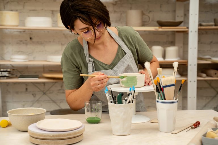 Dark-haired young woman wearing a green shirt and a gray apron and painting pottery with green paint.