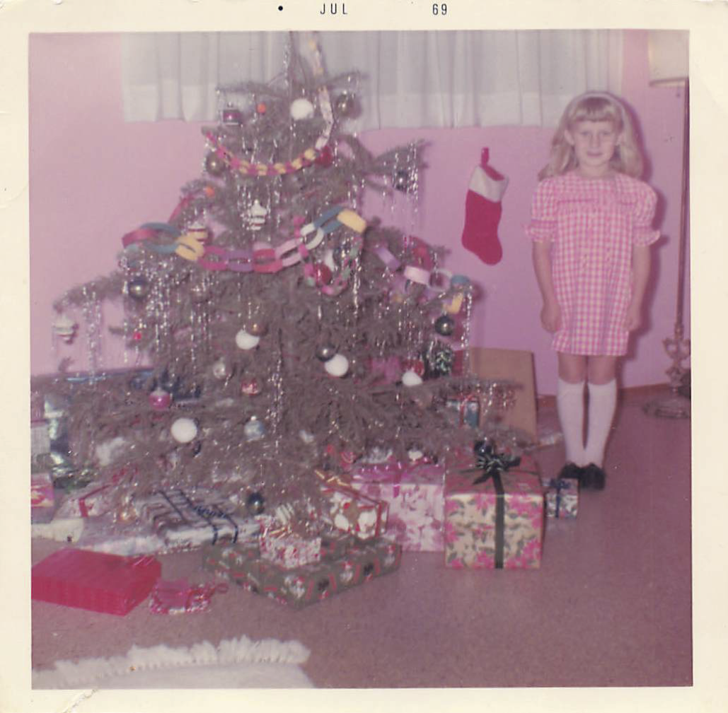 Suzy wearing a pink and white checked dress and white socks standing in a pink and white bedroom next to a real (ugly) Christmas tree with wrapped presents underneath,